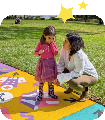 An adult stands next to a little girl playing or telling stories. On the path are brightly coloured illustrations. 