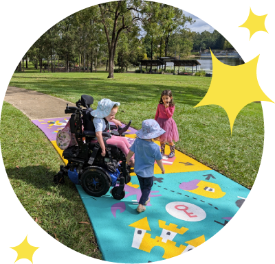 3 girls play on a brightly colour path. One girl is in a wheelchair. The path has a castle, a key, a chest, a shop and other play elements.