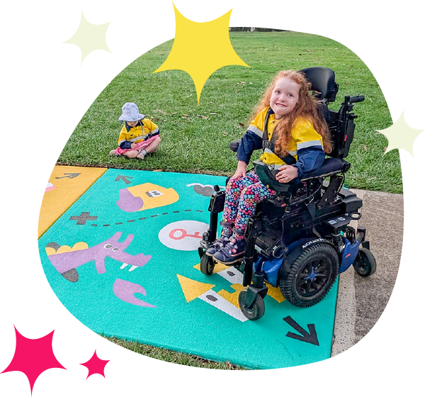 A girl in a wheelchair sits on a colourful painted pavement mat with imaginary play illustrations including a key, castle, and purple dragon. Another child sits nearby on the grass. The play space is outdoors in a park.