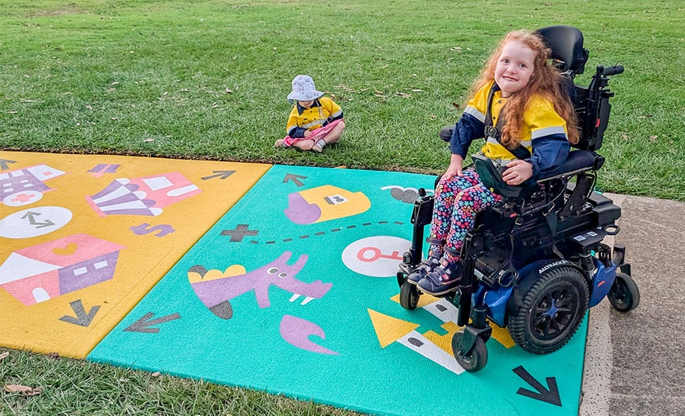 A girl in a wheelchair sits on a colourful painted pavement mat with imaginary play illustrations including a key, castle, and purple dragon. Another child sits nearby on the grass. The play space is outdoors in a park.